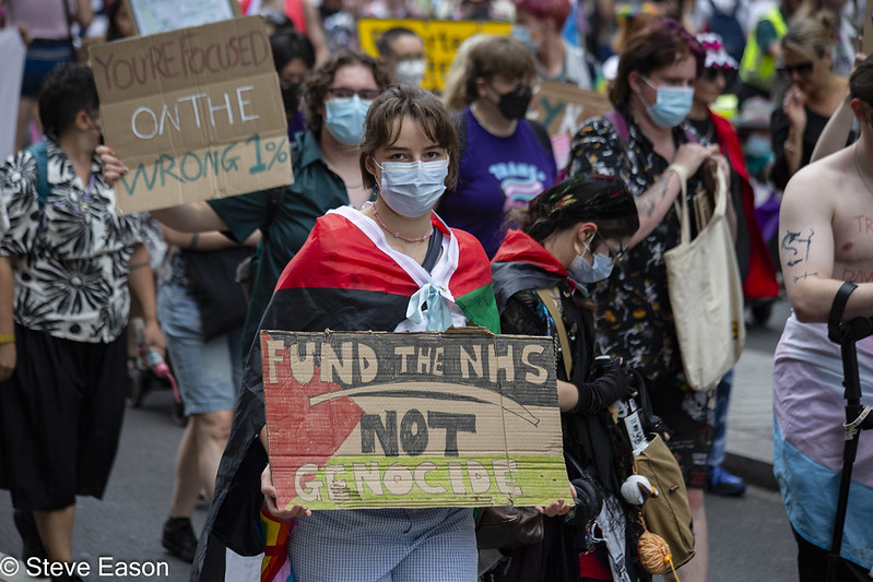 Marchers hold home-made placards reading "You're focused on the wrong 1%" and "Fund the NHS not genocide" over a Palestine flag.