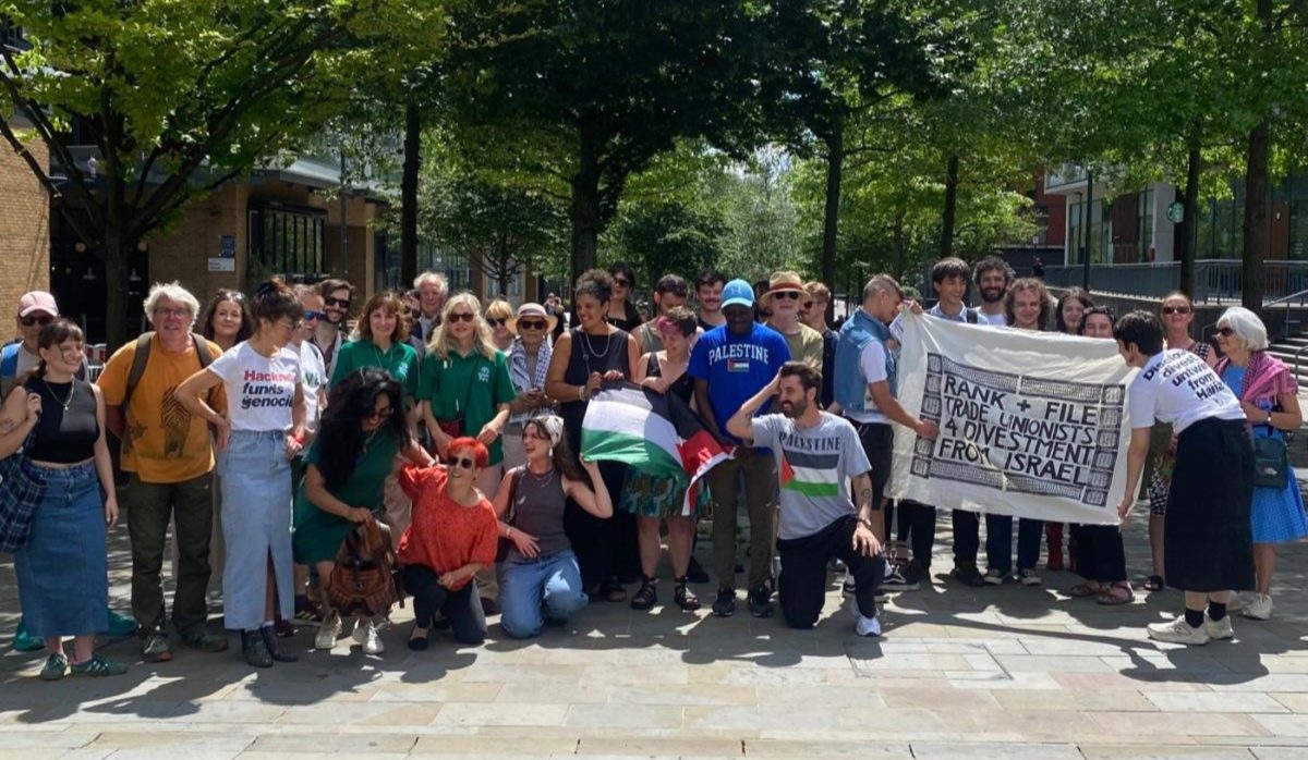 An image of a group of campaigners of "Hackney Votes Palestine" taking a group photo. Many of them are wearing Palestine-related clothes and holding flags of banners. They look happy and are smiling. It's sunny and there's trees in the background.