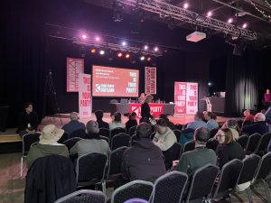 The image is taken by an audience member of the Your Party Sctoland founding conference in Dundee. Audience memebrs are sitting on chairs in severeal rows, looking at the panel in front. Your Party merch banners fill the stage.