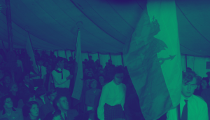 Edit version of a photo of the Capel Celyn Calvinistic Methodist chapel and the Plaid Cymru 'Keep Tryweryn' Rally at Bala. A large group of people sit and stand in a tent. The standing people hold Welsh flags. It gets darker towards the back. There is a green filter over the entire photo.