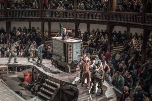 The image shows actors on a stage in front of a theatre audience. They are playing people affected by war, clotehd in rags and with dirty faces. They are pulling Mother Courage's sales wagon.