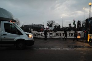 A photo showing a white van and activists blocking an entrance to a factory. They carry a banner saying shut elbit down.