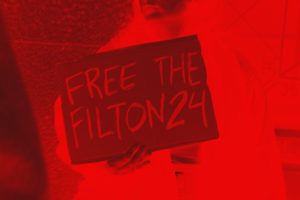 An edited image of a protestor at the UK embassy in Lisbon. They hold a sign saying 'Free the Filton24'. It has been edited to look red with black shadows.