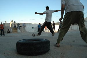 people playing football on sandy ground, with car tires as goal posts. a young man ist jumping towards the ball.
