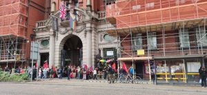 A landscape image of United Kimpton Workers striking outside the Kimpton Clocktower hotel, a red brick building with scaffolding covering it. Red flags can be seen amonst the crowd. On the entrance arch, three flags can be seen - the butcher's apron, a progress pride flag, and a black clocktower flag.