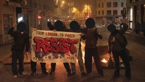 a group of rs21 members with a banner saying "resist, from london to paris" in front of a barricade in the streets of paris